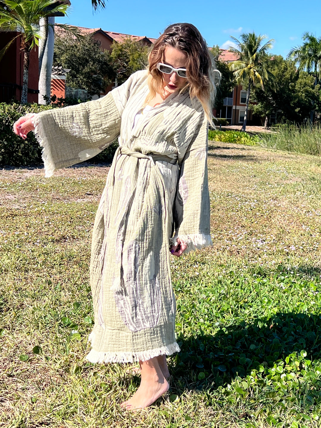 A woman in sunglasses and the Cactus Robe from turkanhome.com stands barefoot on grass, arms outstretched. With sunny palm trees and buildings behind her, this lounge wear doubles as stylish beach wear.