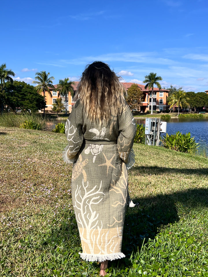A barefoot woman with long, wavy hair stands on grass by a pond, wearing the Green Turtle Robe kimono beach wear from turkanhome.com. Palm trees, water, and colorful buildings appear under a blue sky in the background.