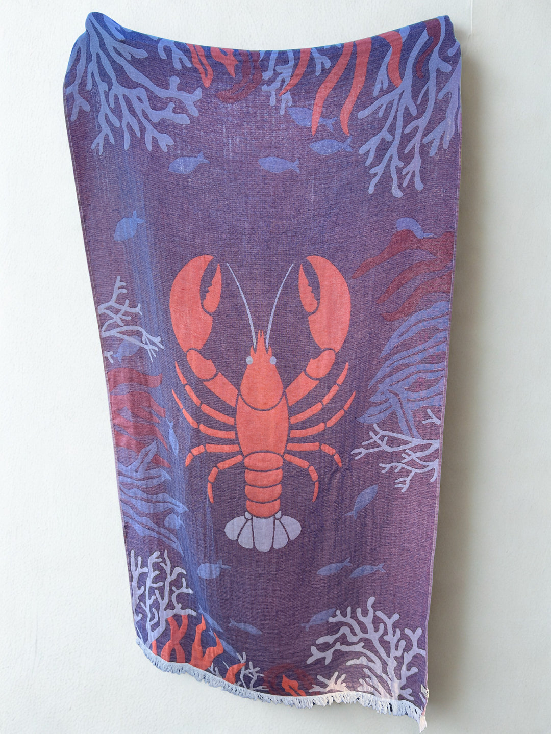 The Lobster Turkish Towel by turkanhome.com is a rectangular, 100% cotton beach towel featuring a large red lobster and blue fish with coral on a purple background, displayed against a white wall.
