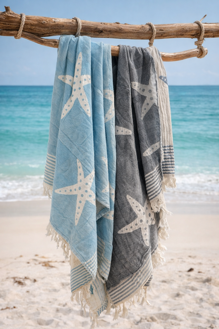 Two Sea Star Turkish Towels by turkanhome.com, Jacquard woven from 100% cotton, hang side by side on a wooden branch with the ocean and sandy beach in the background.