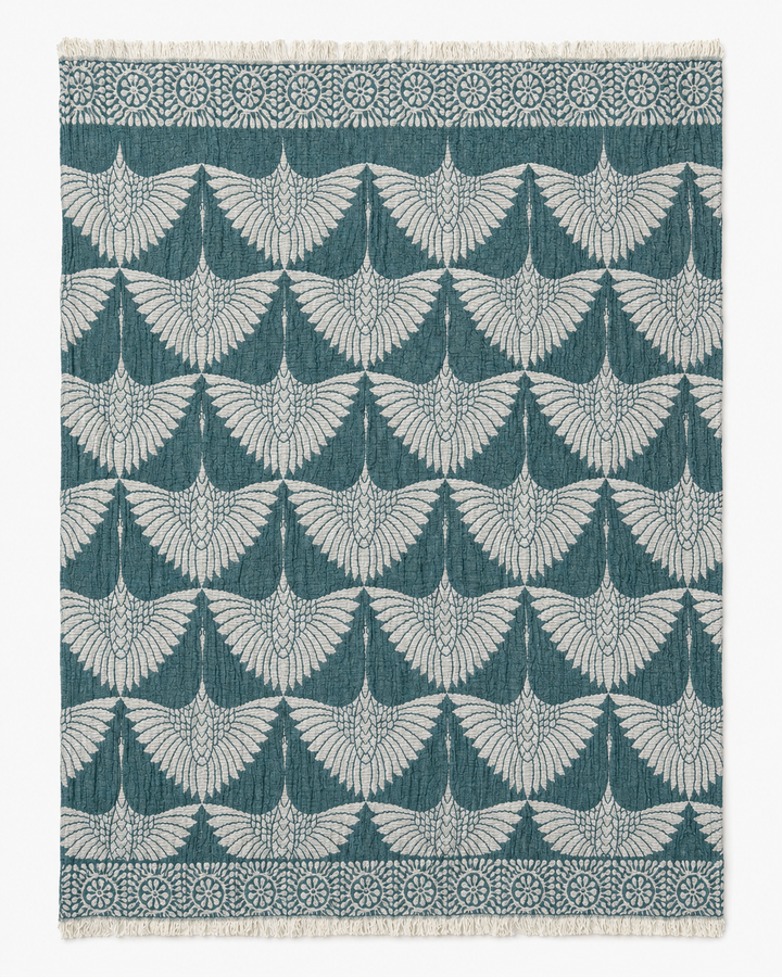 The Crane Muslin Throw Blanket from turkanhome.com features a reversible design with a repeating pattern of stylized crane wings in white on a teal background, accented by a natural decorative border and short fringe on two edges. Crafted from Turkish cotton, this throw offers both style and comfort for your home.