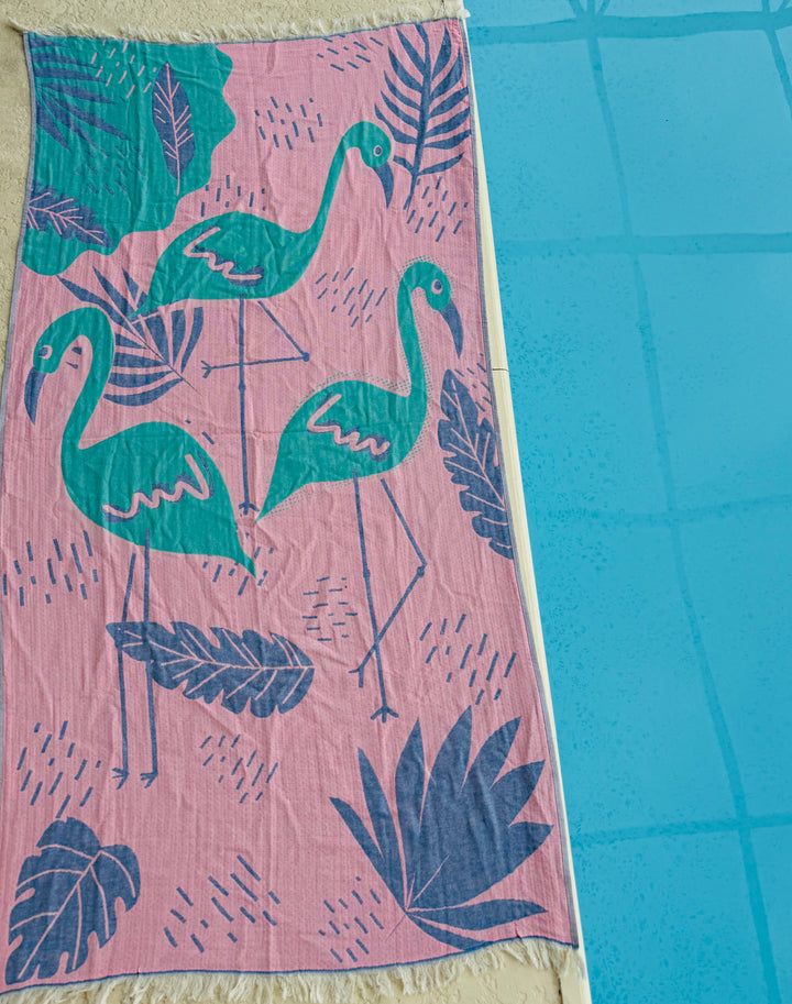 The Flamingo Vibe Towel by turkanhome.com is a quick-drying, 100% cotton beach towel with green flamingos and large leaves on a pink background—perfect for poolside relaxation.
