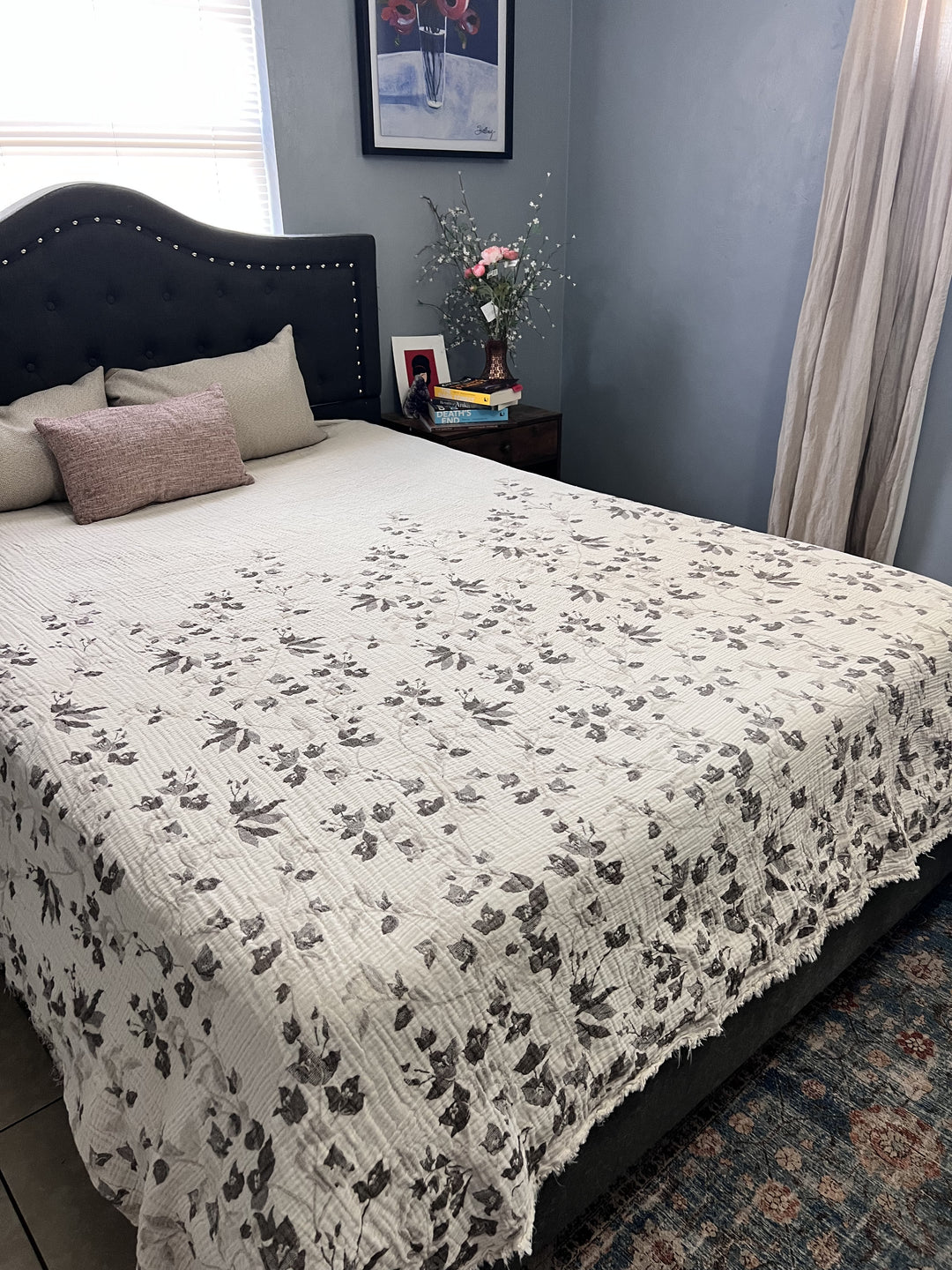 A cozy bedroom features a neatly made bed with a floral quilt, three pillows, brown headboard, and turkanhome.com’s Bougainvillea Muslin Bed Blanket – Brown| Queen. The nightstand displays flowers, books, decor items, and a photo frame.