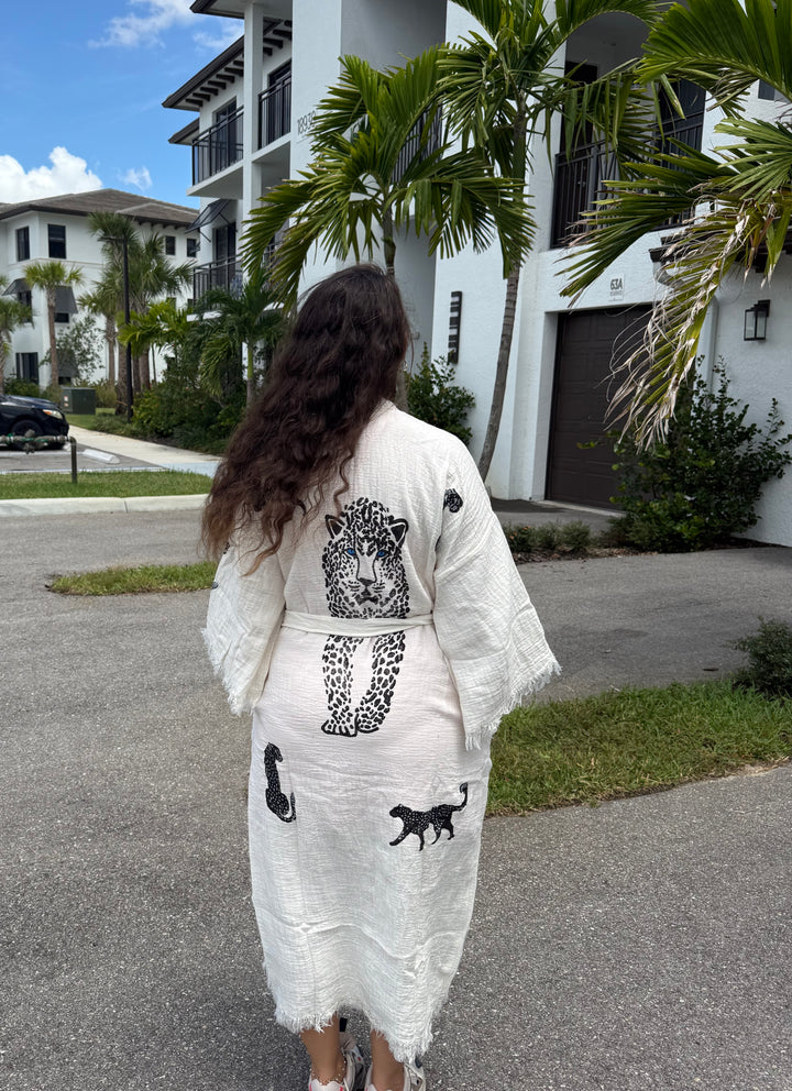 A person with long brown hair, wearing the Leopard Cotton Kimono Robe – Lightweight Boho Beach Cover Up from turkanhome.com, stands on a street near palm trees and white buildings, facing away from the camera.