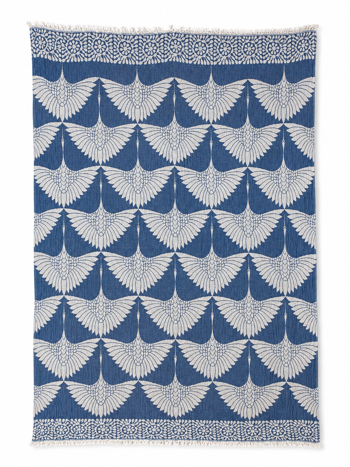 The Crane Muslin Throw Blanket Natural Border by turkanhome.com features a bold, reversible design with a striking blue base and a repeating white crane motif, each bird depicted with outstretched wings. Crafted from premium Turkish muslin cotton, this throw is accented by a perfectly symmetrical geometric floral border for added elegance.