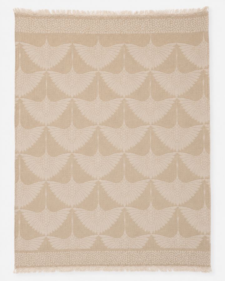 The Crane Muslin Throw Blanket in Natural Border from turkanhome.com features a beige Turkish cotton textile adorned with a repeating motif of stylized cranes in flight, wings outstretched, and is finished with decorative borders at the top and bottom edges. Expertly crafted as a reversible throw, it includes fringe along the shorter sides.