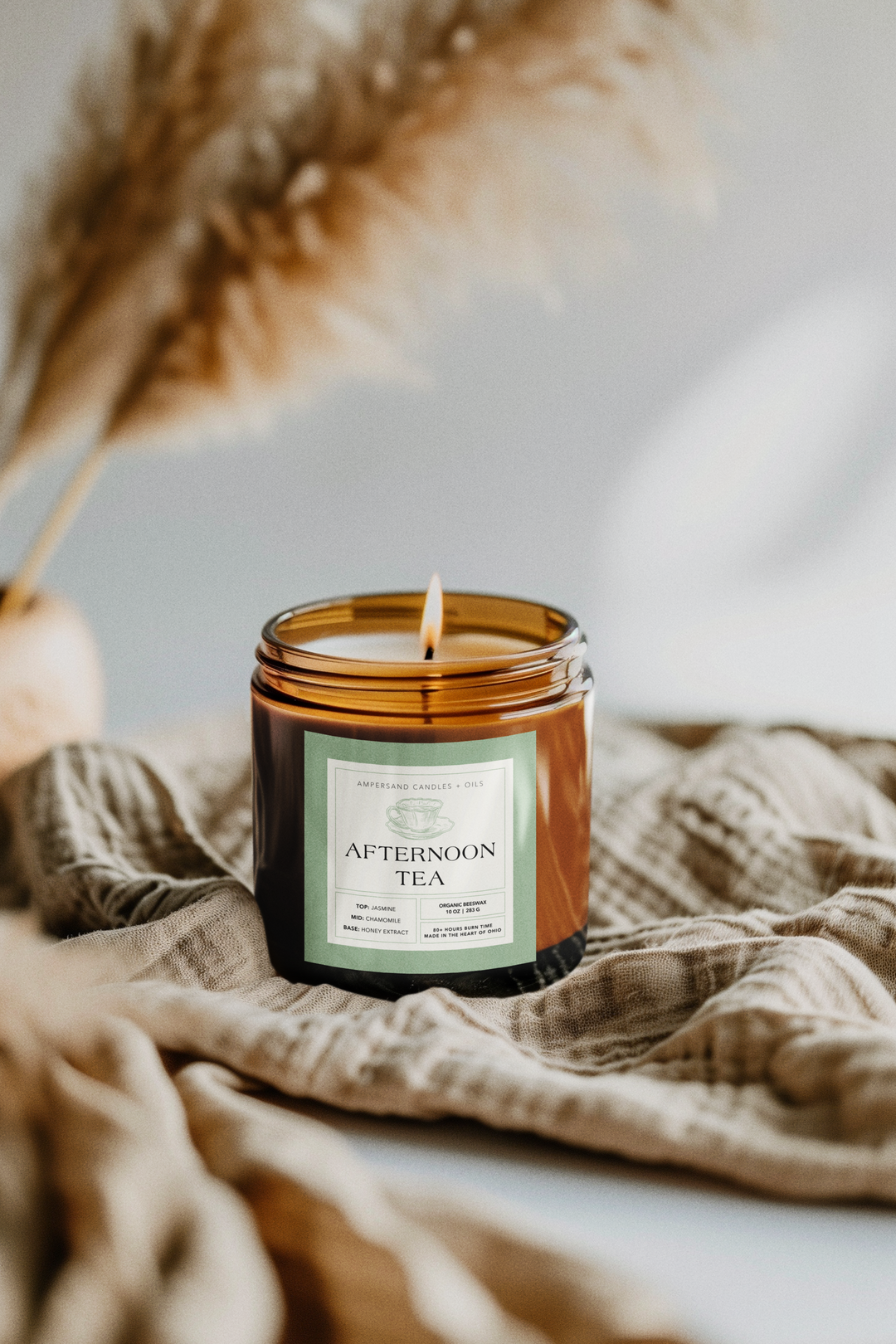 An Ampersand Candles + Oils Afternoon Tea Beeswax Candle (10oz) rests on soft beige fabric, while blurred pampas grass in the background adds to the candles cozy, relaxing ambiance.