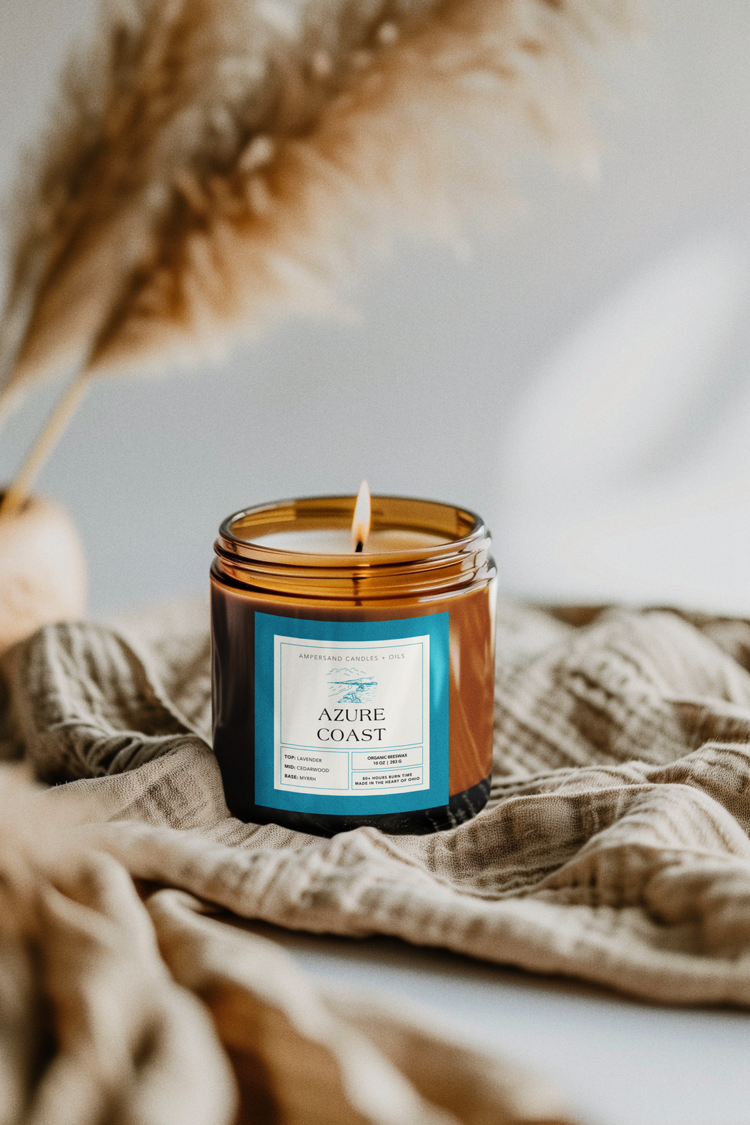 A lit Ampersand Candles + Oils Azure Coast Beeswax Candle (10oz) rests on a beige textured cloth, with soft-focus pampas grass behind it, creating a cozy, calming beach-inspired vibe.