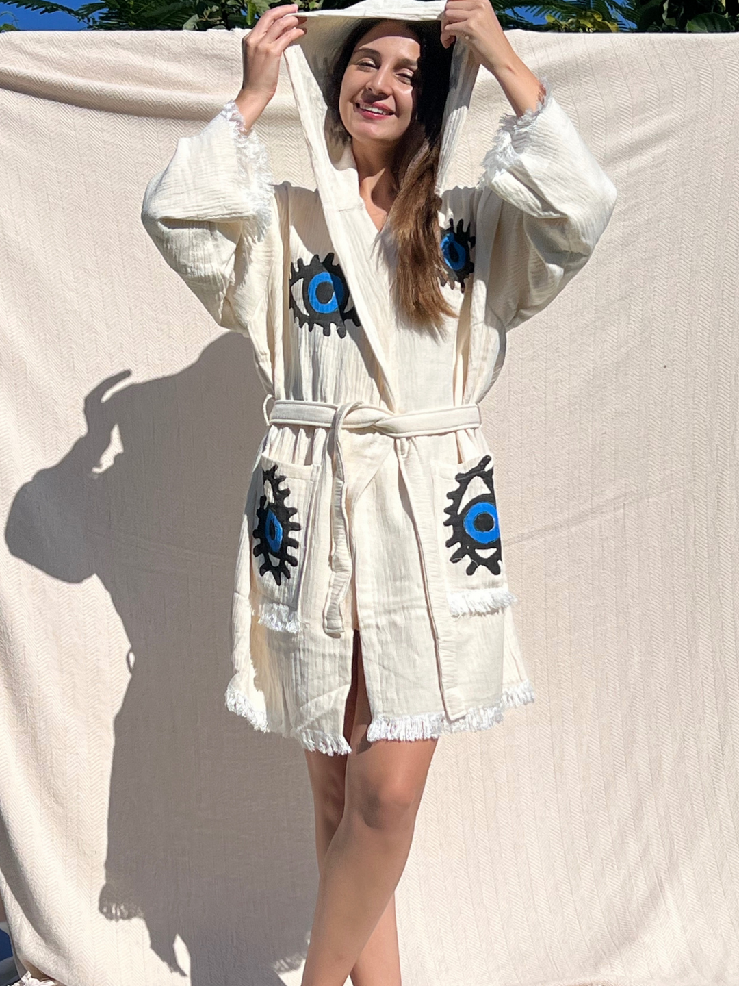 A smiling woman poses outdoors in front of a beige backdrop, wearing the Blue Eye Robe—Short with Pockets and Hood by turkanhome.com, featuring blue and black eye designs, perfect for lounge or beach wear.
