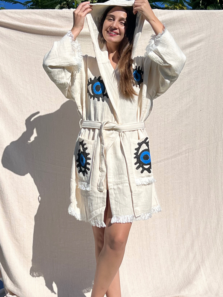 A smiling woman poses outdoors in front of a beige backdrop, wearing the Blue Eye Robe—Short with Pockets and Hood by turkanhome.com, featuring blue and black eye designs, perfect for lounge or beach wear.