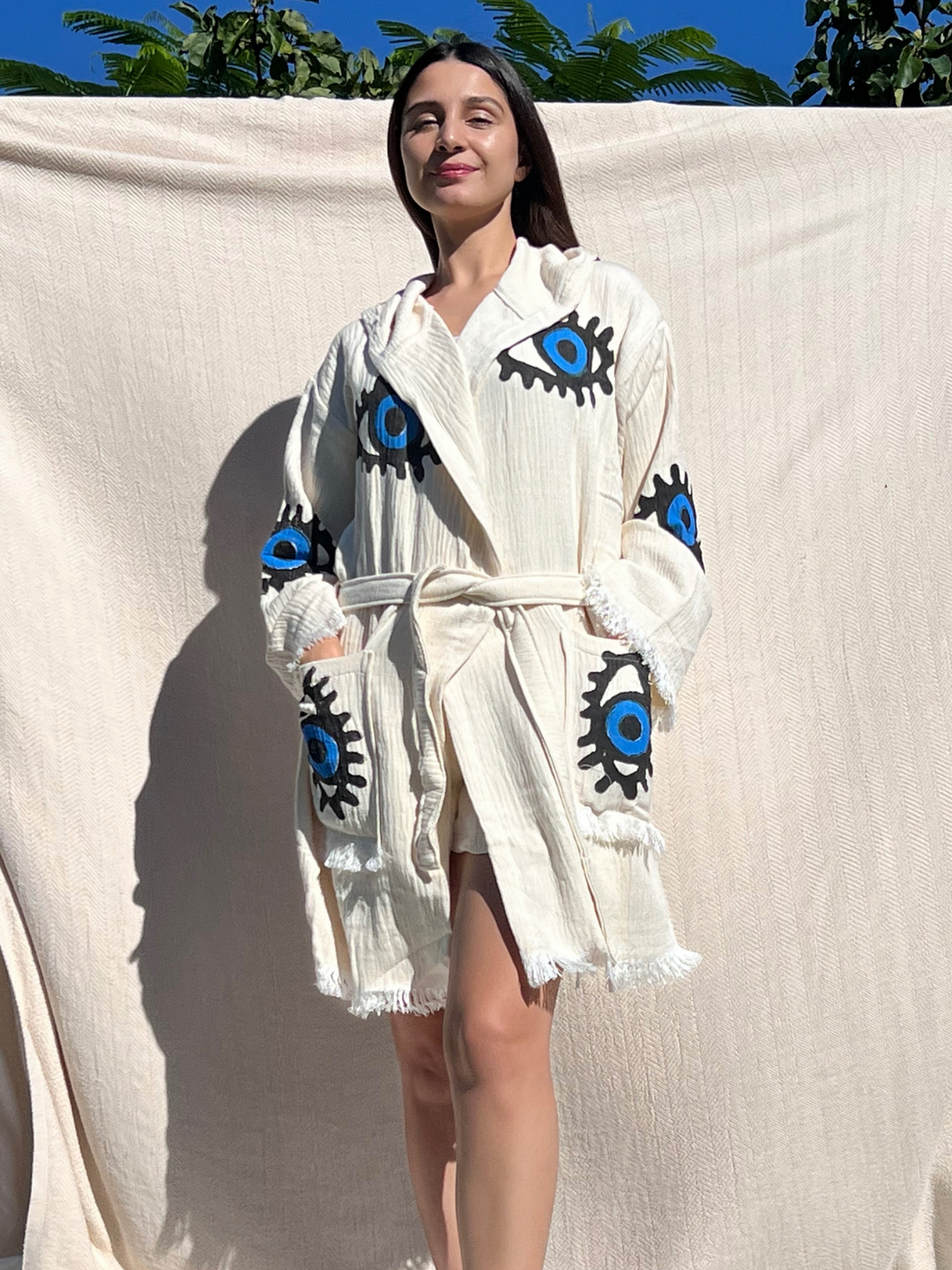A woman stands outdoors in sunlight wearing the Blue Eye Robe–Short with Pockets and Hood from turkanhome.com, its belted boho kimono style showing bold blue and black eye designs, surrounded by a cream cloth, green leaves, and blue sky.