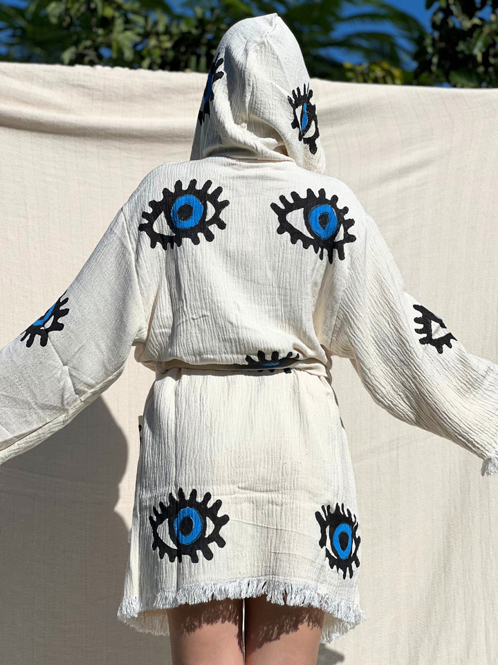 A person stands with arms outstretched, wearing turkanhome.com's Blue Eye Robe—short, cream-colored cotton lounge or beach wear with pockets, hood, and bold blue and black eye designs. Background features neutral fabric, greenery, and sky.