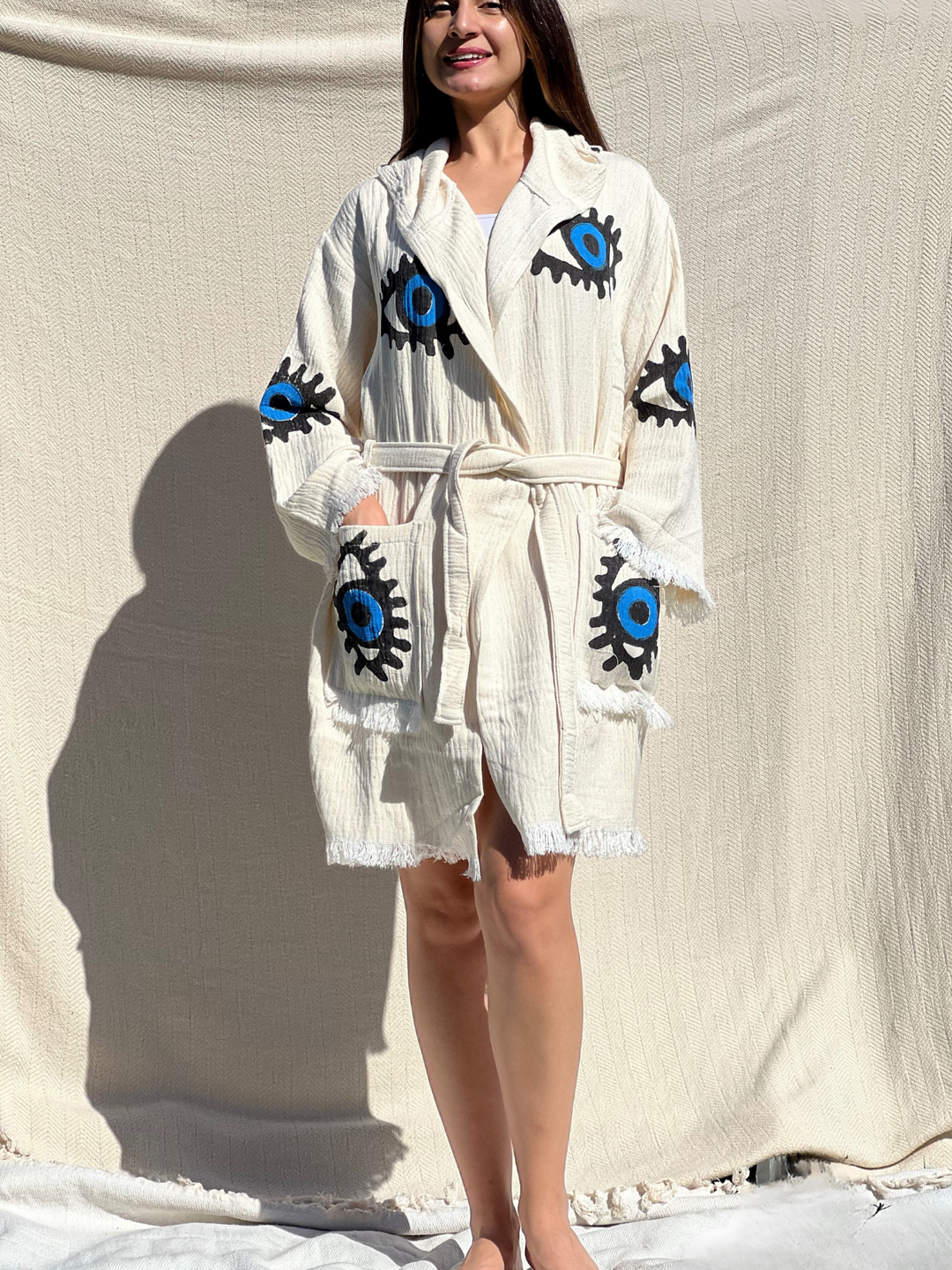 A woman poses before a light backdrop in the Blue Eye Robe—Short with Pockets and Hood by turkanhome.com, a cream cotton kimono-style lounge and beach wear featuring bold blue and black eye motifs on the chest, sleeves, and pockets.