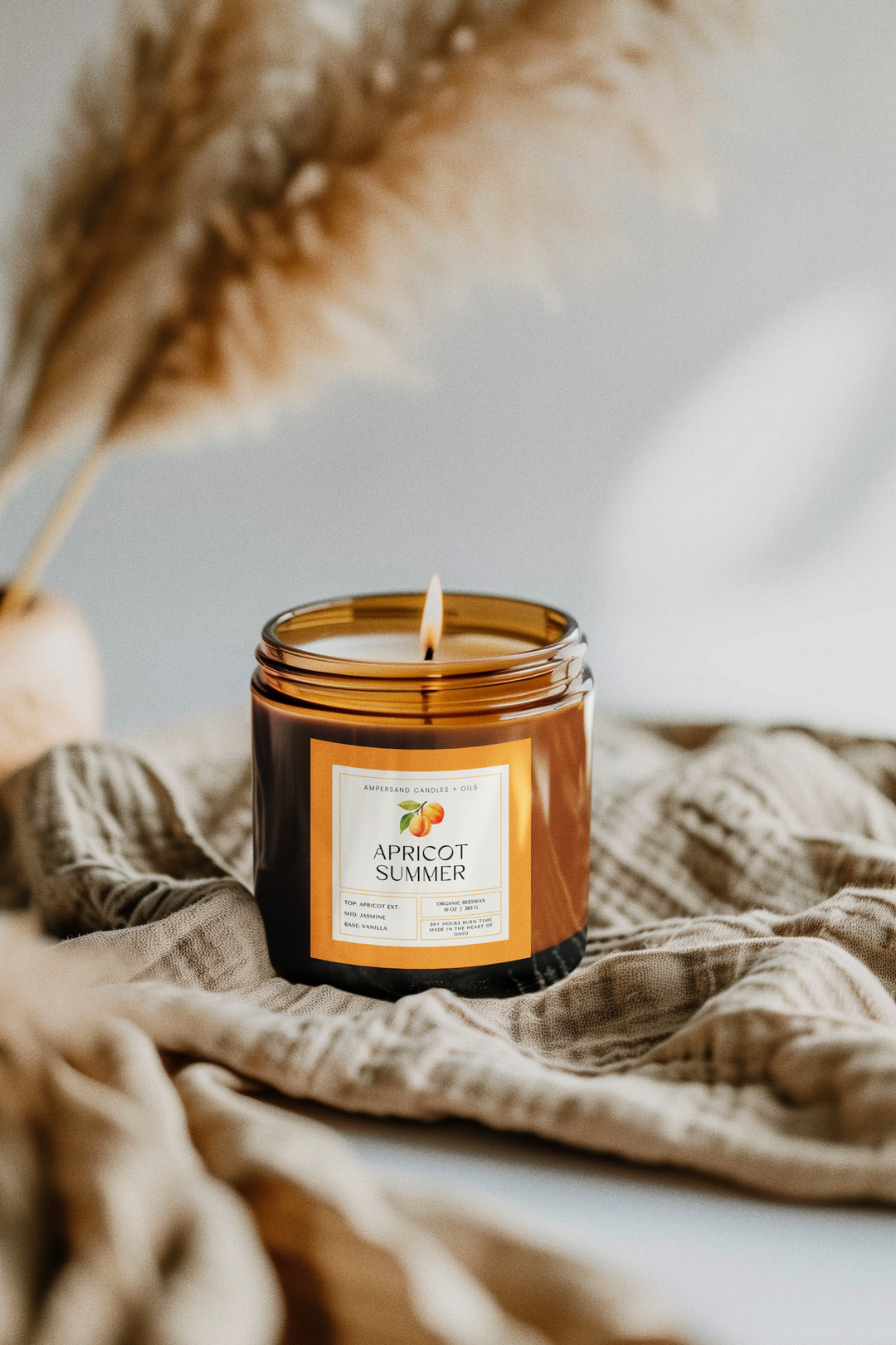The Apricot Summer Beeswax Candle (10oz) by Ampersand Candles + Oils glows in a brown glass jar atop a beige textured cloth, with blurred dried plants behind it, creating a cozy and inviting atmosphere.