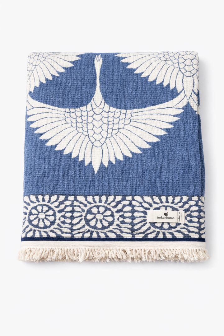 The Crane Muslin Throw Blanket – Reversible Turkish Cotton by turkanhome.com features a blue and white crane design with floral patterns, soft muslin cotton, fringe edges, and a branded label.