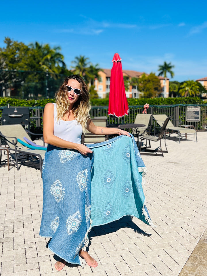 A woman in sunglasses stands by a pool holding the turkanhome.com Eye Blue Sand Resistant Turkish Beach Towel, Throw, Shawl. Lounge chairs, a closed red umbrella, palm trees, and blue sky are visible in the background.