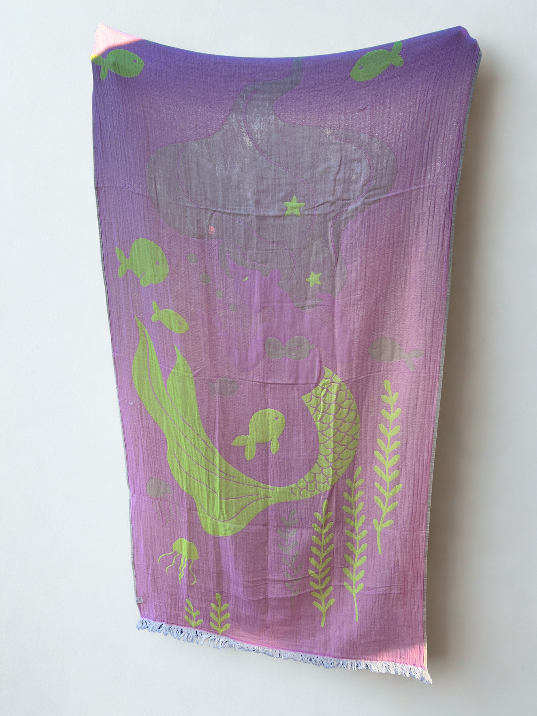 The Mermaid Turkish Towel from turkanhome.com is a purple and pink 100% cotton beach towel with green and gray mermaid, fish, seaweed, and bubble illustrations, shown flat against a light background.