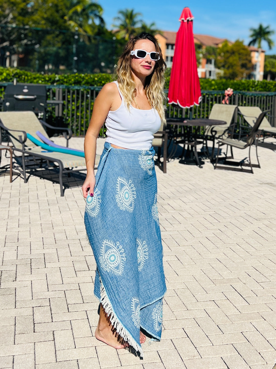 A woman in a white tank top, blue wrap skirt, and white sunglasses stands barefoot by a sunlit poolside with lounge chairs, a red umbrella, and the Eye Blue Sand Resistant Turkish Beach Towel by turkanhome.com in the background.