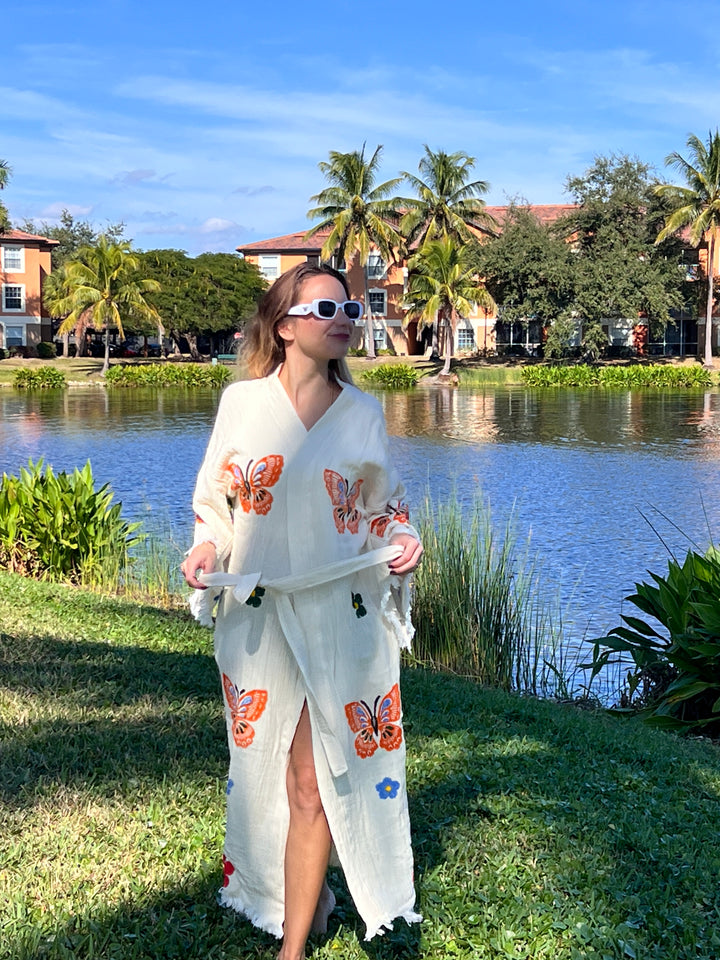 A woman in sunglasses wears the Butterfly Kimono Robe-Natural Color from turkanhome.com—a white dress with orange butterfly prints—while standing on grass beside a lake, with palm trees and buildings under a blue sky.