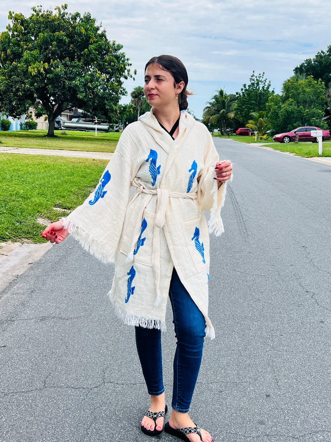 A woman in a turkanhome.com Seahorse Kimono Robe with Hoodie, featuring blue designs, walks barefoot on a suburban street in dark jeans and leopard-print sandals. Trees and houses line the background beneath a cloudy sky.