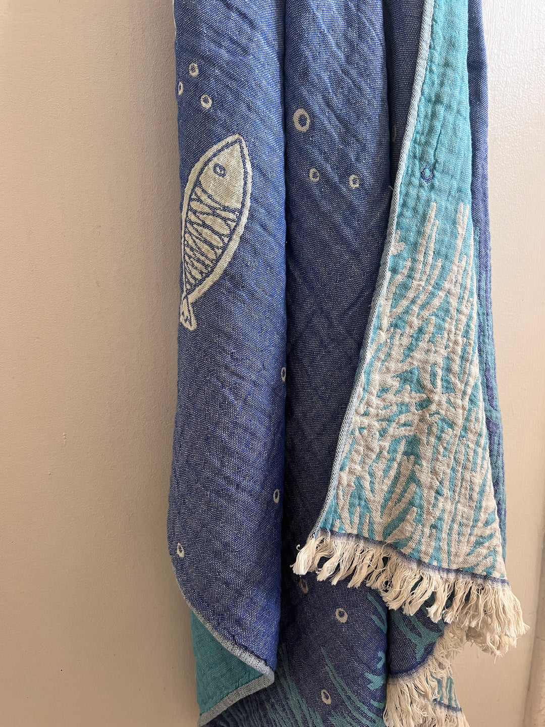 The Fishes and Coral Turkish Towel by turkanhome.com is a blue and teal 100% cotton jacquard with white fish and coral designs, hanging against a white wall. One edge features white fringe for added detail.