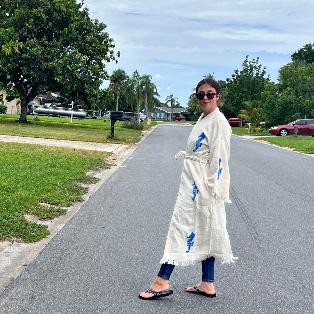 A woman stands on a quiet suburban street, smiling back in sunglasses, jeans, sandals, and the Seahorse Kimono Robe by turkanhome.com. Green lawns, trees, and parked cars complete the peaceful neighborhood scene.