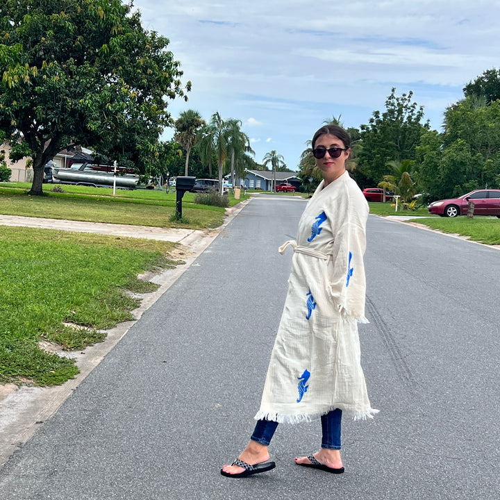 A woman stands on a quiet suburban street, smiling back in sunglasses, jeans, sandals, and the Seahorse Kimono Robe by turkanhome.com. Green lawns, trees, and parked cars complete the peaceful neighborhood scene.