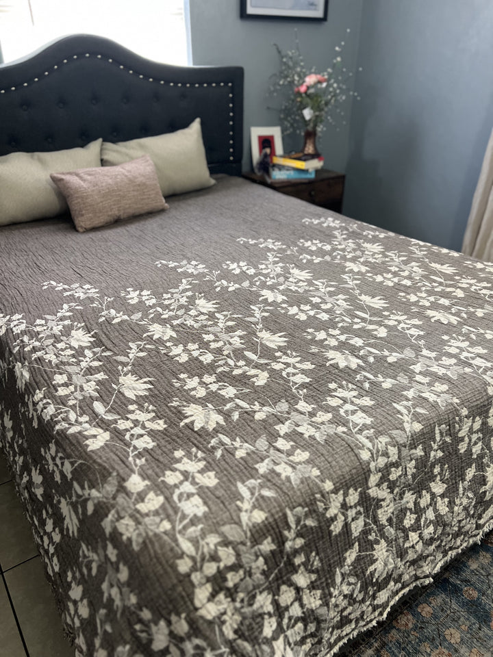 A neatly made bed with a dark tufted headboard, light pillows, and the turkanhome.com Bougainvillea Muslin Bed Blanket – Brown| Queen, featuring a white floral pattern. A wooden bedside table holds a framed photo, figurine, and vase with flowers.