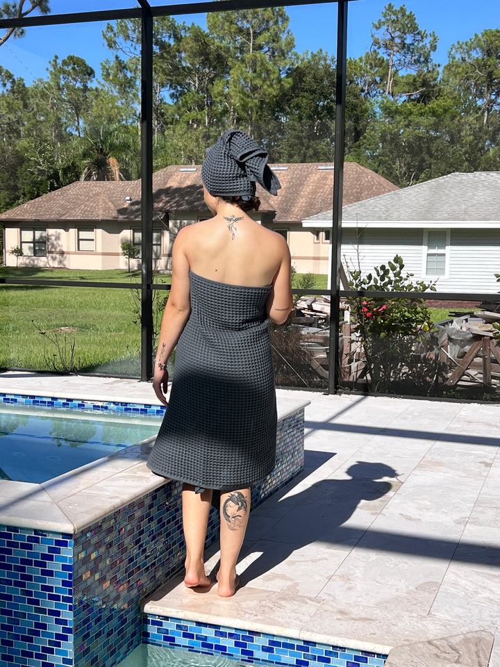 A person stands barefoot by a pool, wrapped in a turkanhome.com Waffle Bath Towel. Outdoors, greenery, trees, and houses can be seen in the background.