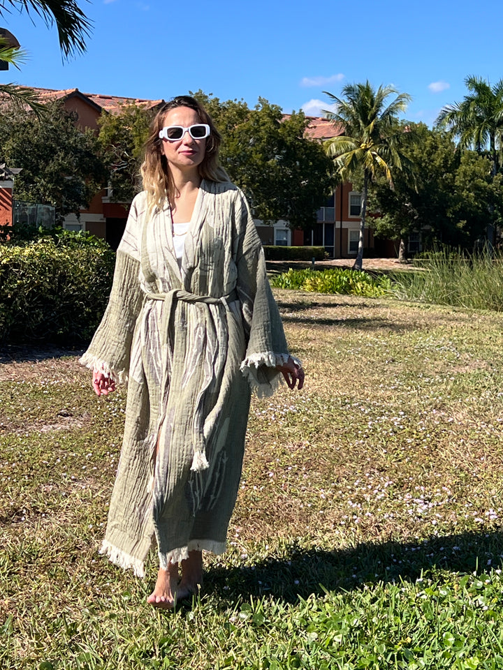 A woman in a Cactus Robe from turkanhome.com, styled as lounge or beach wear, walks barefoot on sunny grass with palm trees and colorful buildings behind her, wearing white sunglasses.