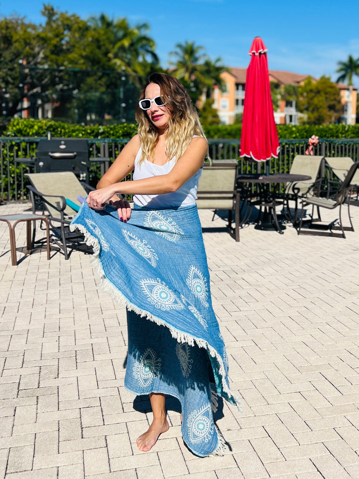 A woman in sunglasses and a white tank top wraps the Eye Blue Sand Resistant Turkish Beach Towel by turkanhome.com around her waist while standing barefoot on a sunny patio with chairs, tables, and a red umbrella.