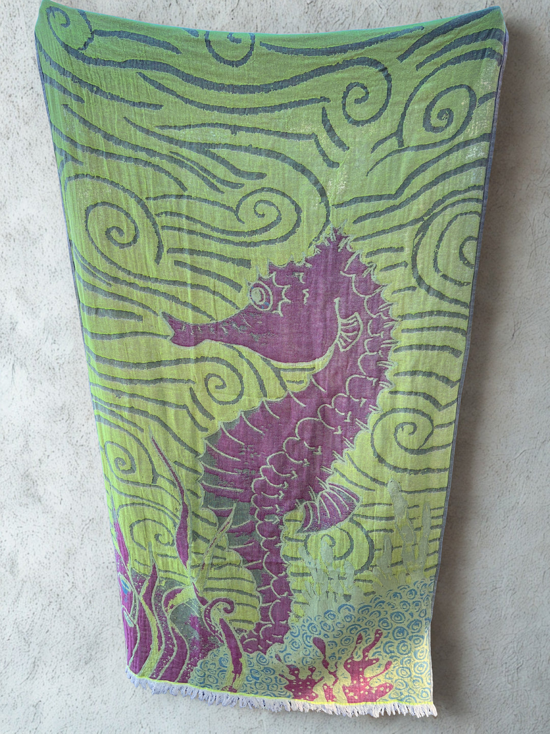 The Seahorse Turkish Towel from turkanhome.com, made of 100% cotton, features a purple seahorse, swirling green patterns, and red-green seaweed accents. It hangs on a light beige wall and offers quick-drying performance.