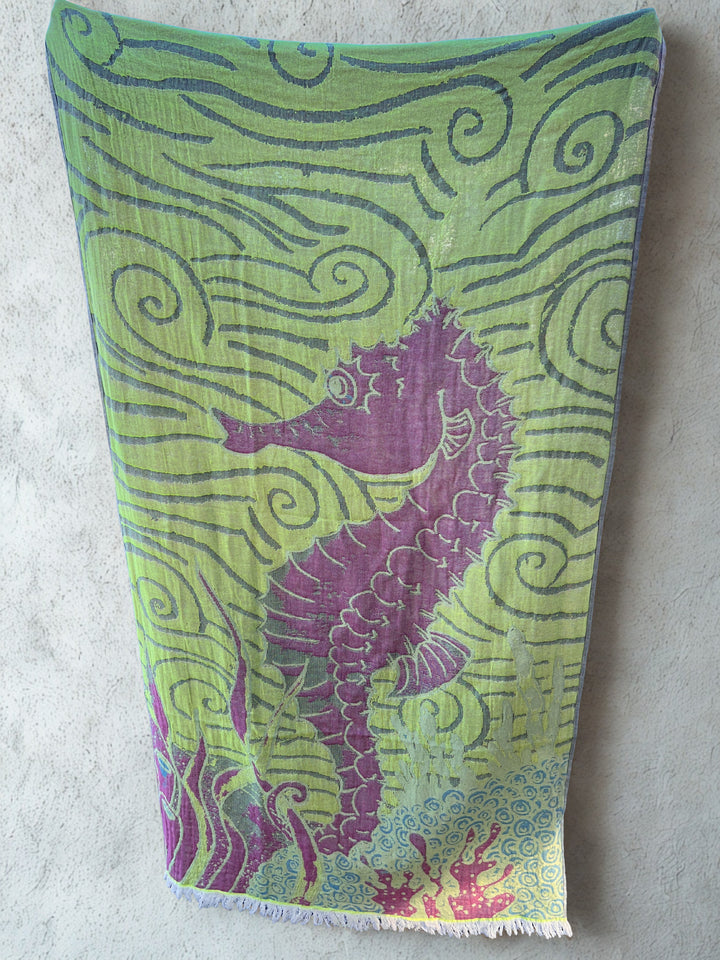 The Seahorse Turkish Towel from turkanhome.com, made of 100% cotton, features a purple seahorse, swirling green patterns, and red-green seaweed accents. It hangs on a light beige wall and offers quick-drying performance.