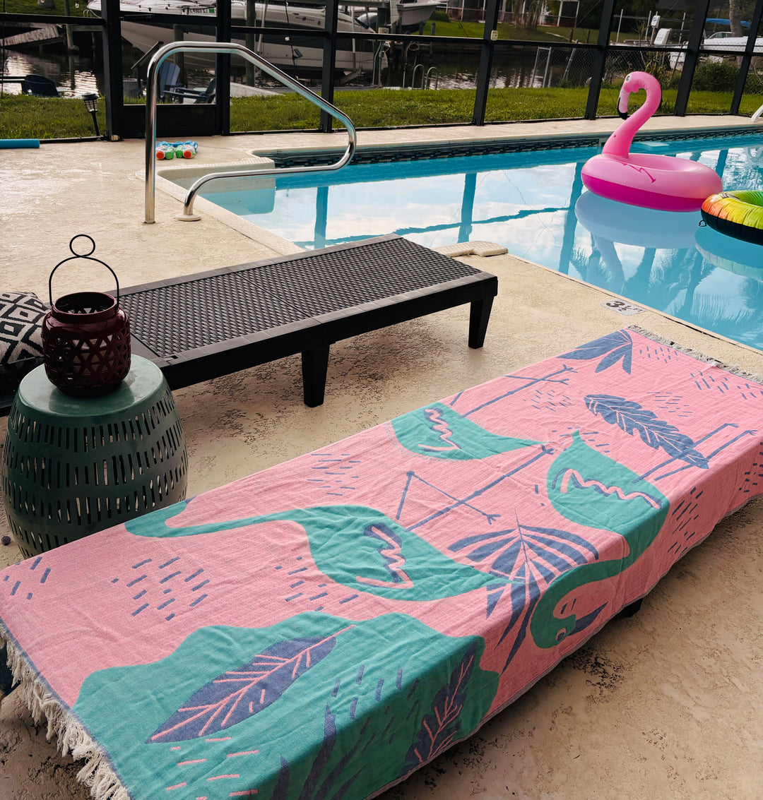 The Flamingo Vibe Towel – Quick Drying Towel from turkanhome.com, featuring green leaf patterns, covers a lounge chair near the pool and pink flamingo float. A black bench, lantern, cushion, green stool, and canal boats are nearby.