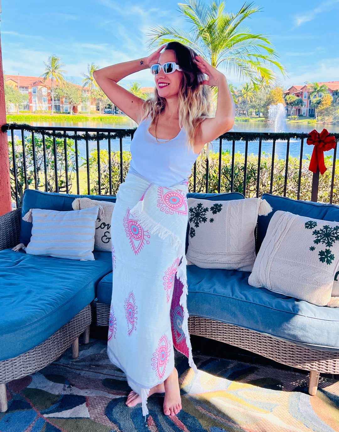 A woman in sunglasses and a white tank top stands barefoot on an outdoor patio, smiling with hands in her hair. She wears the Eye Red Sand Resistant Turkish Beach Towel by turkanhome.com, surrounded by blue seats, plants, and a lake.