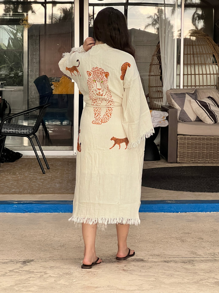A woman stands with her back to the camera, wearing the Orange Leopard Cotton Kimono Robe from turkanhome.com—a lightweight boho beach cover up. She faces a glass door, with patio furniture and plants visible inside.
