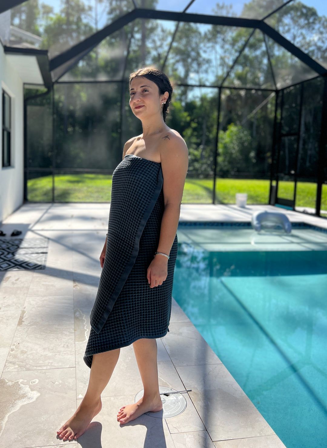 A person wrapped in a dark-checkered Waffle Bath Towel from turkanhome.com stands barefoot by a sunlit pool, surrounded by greenery and a screened enclosure.