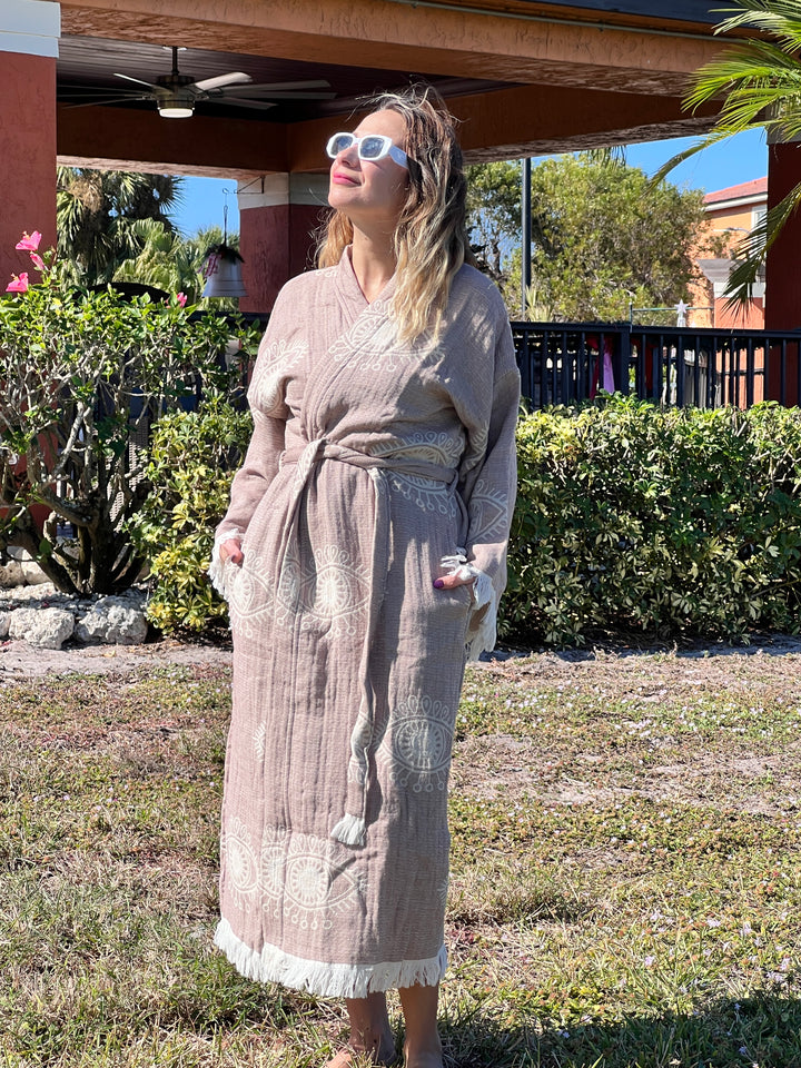 A woman in a Brown Eye Robe Kimono by turkanhome.com stands on grass outdoors, smiling and looking up with sunglasses, surrounded by greenery, flowers, and a building under the sun.