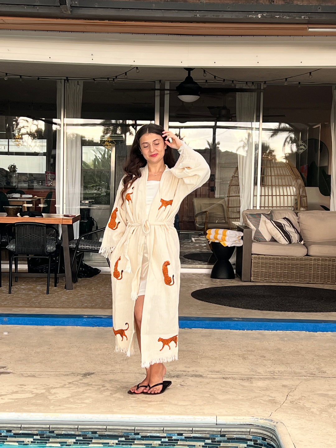 A woman with long dark hair stands by a pool, wearing the turkanhome.com Orange Leopard Cotton Kimono Robe as a lightweight boho beach cover up, paired with a white dress and black flip-flops. Outdoor patio furniture is in the background.