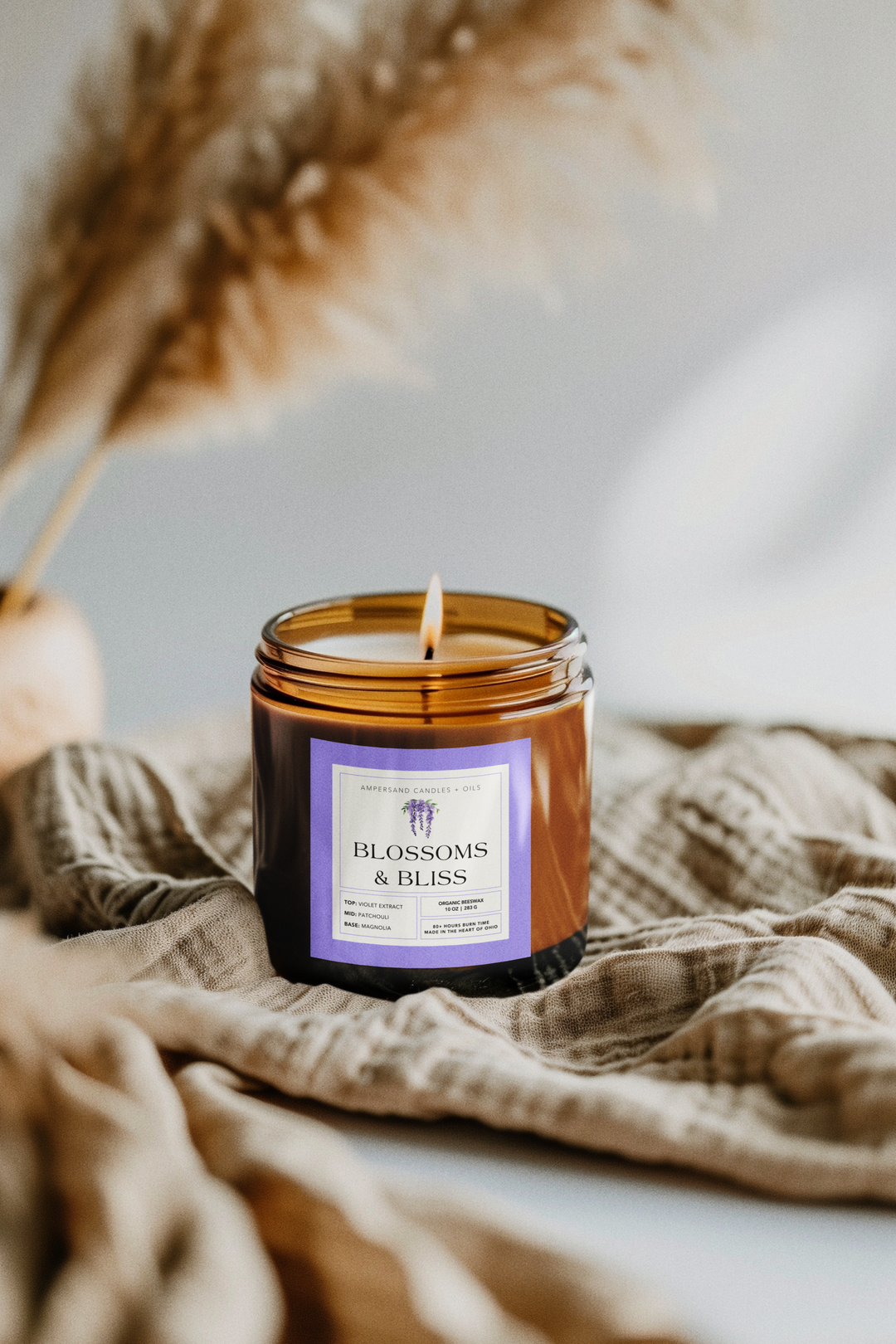 A lit Blossoms & Bliss Beeswax Candle (10oz) by Ampersand Candles + Oils, made from organic beeswax, sits on a beige fabric with soft-focus pampas grass in the background.