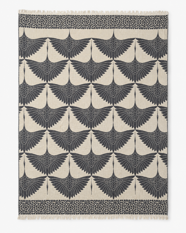 Rewritten Sentence: The Crane Muslin Throw Blanket Natural Border by turkanhome.com is a rectangular, reversible Turkish cotton throw featuring a distinctive crane motif with repeating stylized black birds in flight on a cream background. Decorative black borders adorned with circular and floral accents along the top and bottom edges evoke traditional Turkish artistry.