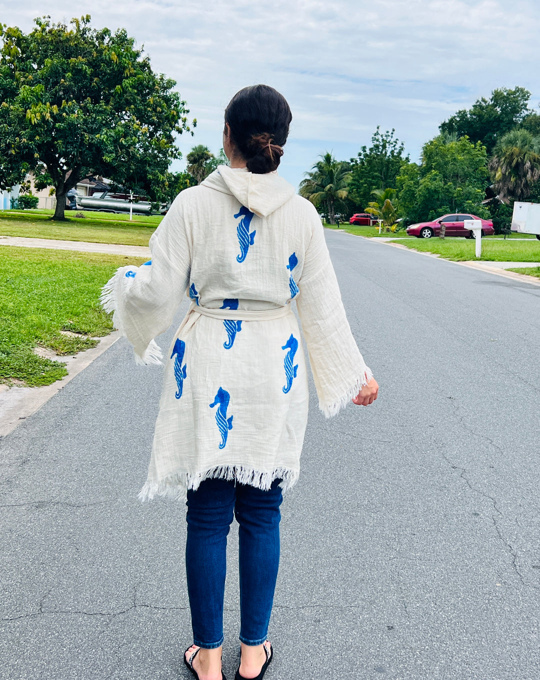 A person with dark hair in a low bun wears the turkanhome.com Seahorse Kimono Robe with Hoodie—a white, eco-friendly robe featuring blue seahorses—paired with dark jeans and sandals, standing on a tree-lined suburban street.