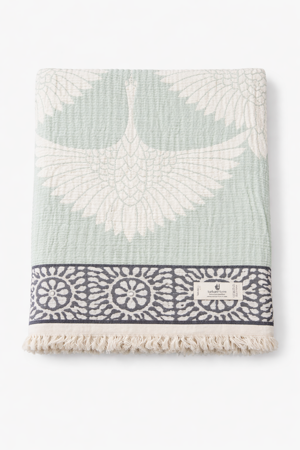 The Crane Muslin Throw Blanket – Reversible Turkish Cotton by turkanhome.com features a white crane on light green, navy and white floral border, fringe edges, sewn-on label, and is crafted from soft Turkish muslin cotton for comfort and style.