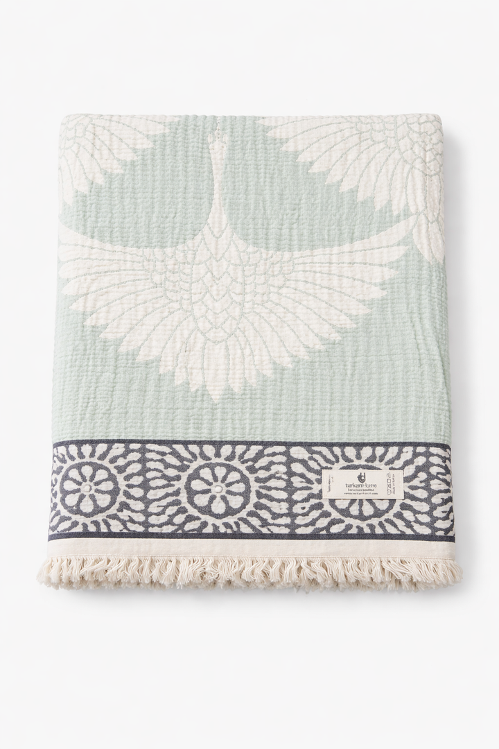 The Crane Muslin Throw Blanket – Reversible Turkish Cotton by turkanhome.com features a white crane on light green, navy and white floral border, fringe edges, sewn-on label, and is crafted from soft Turkish muslin cotton for comfort and style.