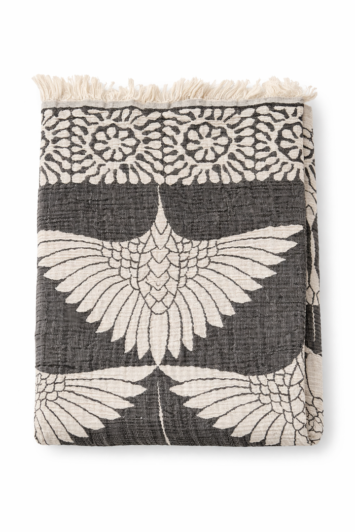 A folded black Crane Muslin Throw Blanket – Reversible Turkish Cotton by turkanhome.com features a white bird and floral pattern with a fringe border.