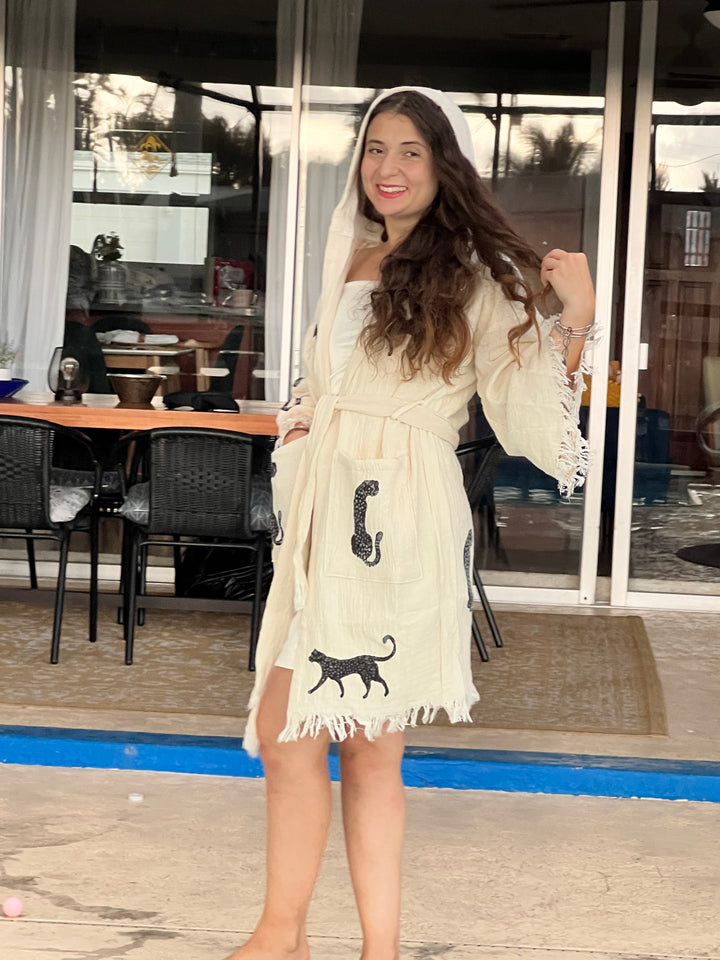 A smiling woman with long hair wears the Black Leopard Hooded Cotton Kimono Robe by turkanhome.com. She stands barefoot outdoors before a glass patio door, with indoor table and chairs visible in the background.