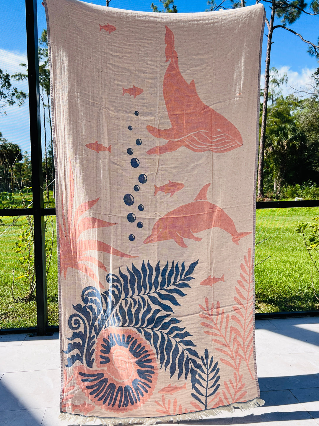 The Dolphin and Whale Turkish Towel by turkanhome.com, with jacquard-woven pink sea life designs, including dolphins and whales, is displayed outdoors in front of a green lawn and trees on a sunny day.