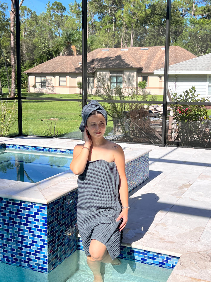 A woman outdoors near a house relaxes by a small pool, wearing a gray Waffle Hand Towel from turkanhome.com and a matching head wrap. One leg dips in the water as she touches her face; trees and lawn are visible in the background.