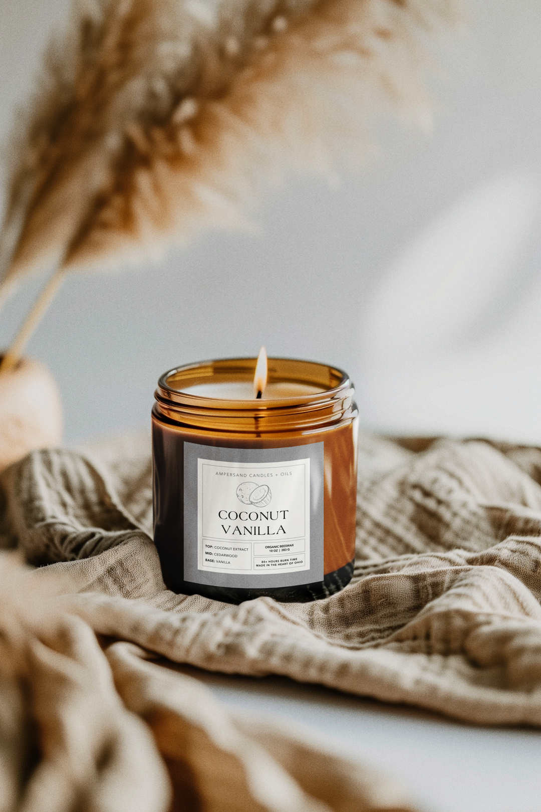 A lit Ampersand Candles + Oils Coconut Vanilla Beeswax Candle (10oz) glows in a brown glass jar atop a beige cloth, with pampas grass softly blurred in the background, creating a clean burn ambiance.