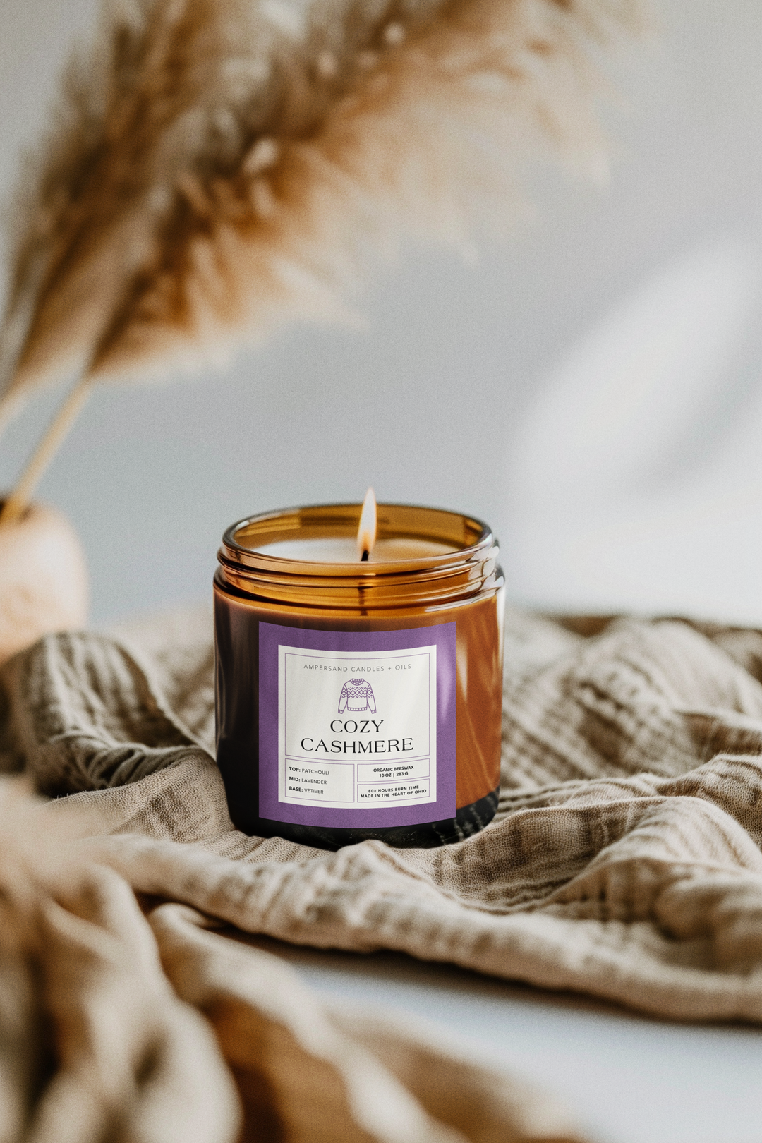A lit Cozy Cashmere Beeswax Candle (10oz) by Ampersand Candles + Oils, made with pure essential oils, sits on soft beige fabric, with dried pampas grass blurred in the background.